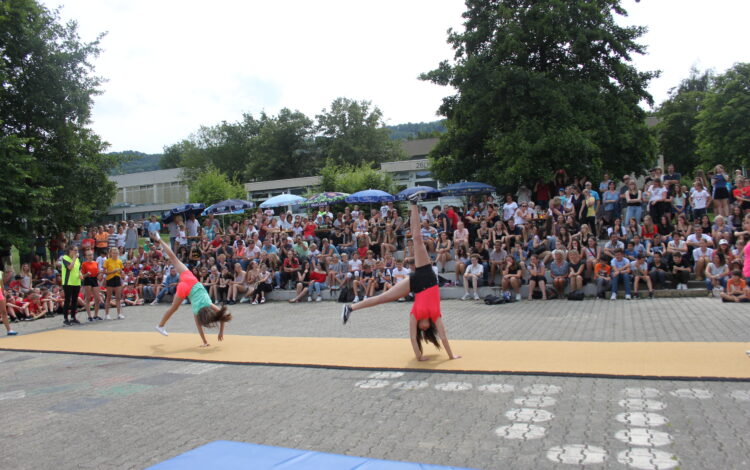 Zwei Schülerinnen schlagen auf einer Turnmatte auf dem Schulhof ein Rad. Im Hintergrund sieht viel Publikum den beiden zu.