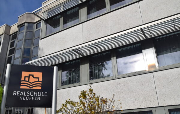 Schild mit dem Logo der Realschule Neuffen vor dem Schulgebäude