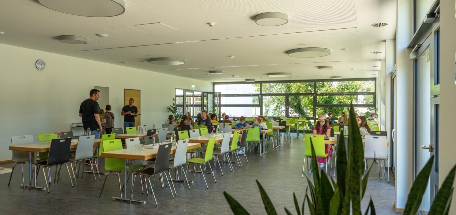 Essraum mit Tischen und Stühle in der Mensa. An ein paar Tischen sitzen Schüler und essen.