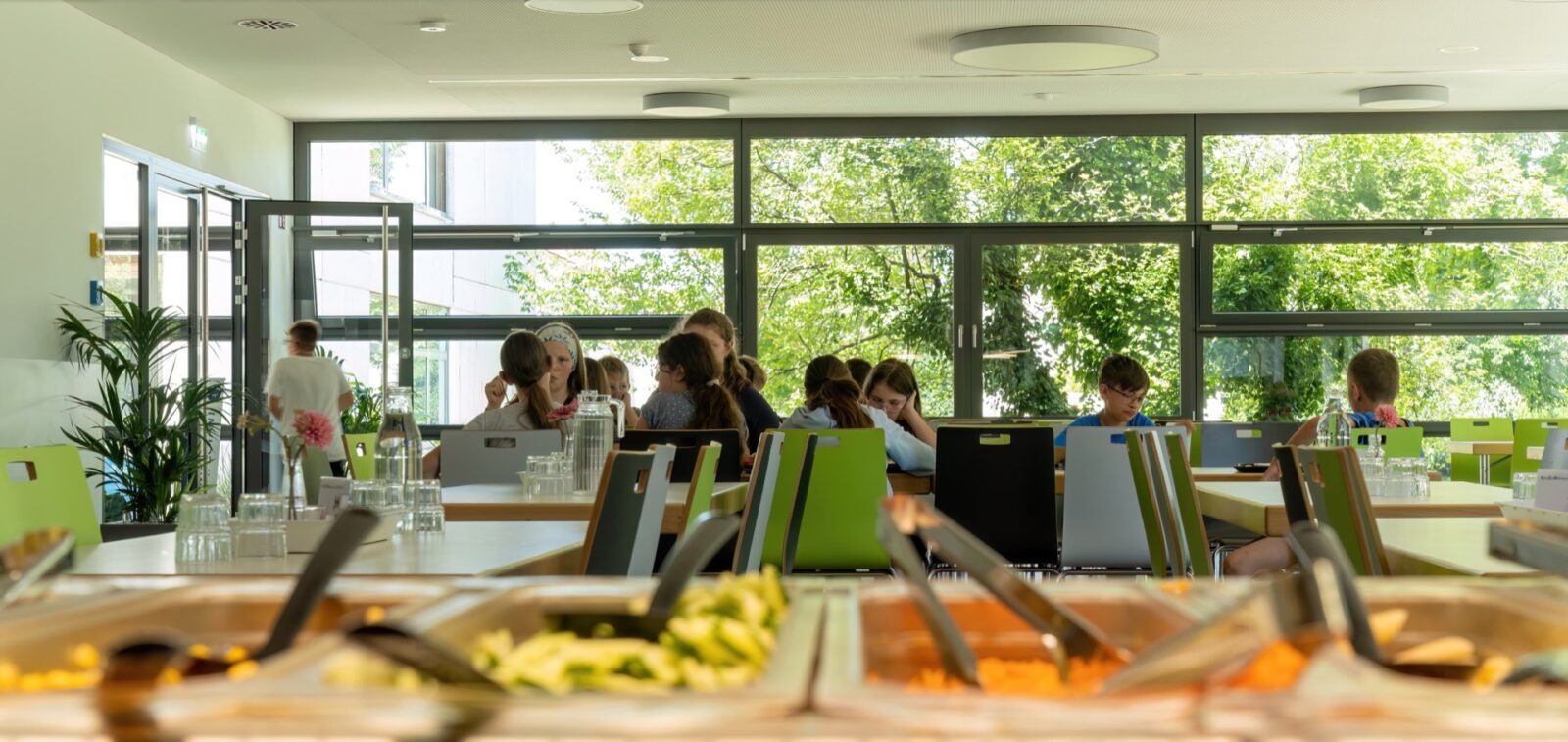 Schüler sitzen an Tischen in der Mensa und essen. Im Vordergrund die Essensausgabe.