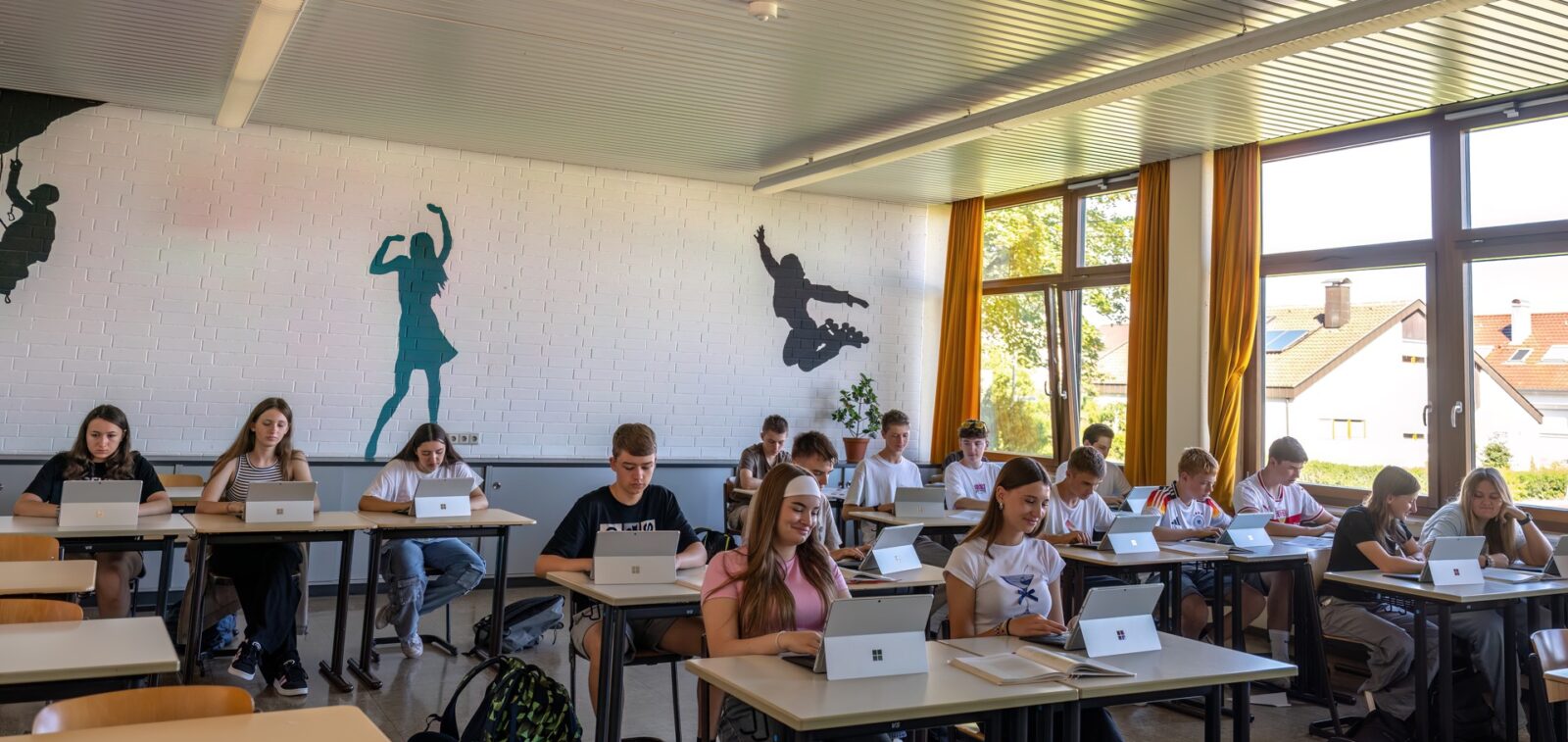 Schülerinnen und Schüler sitzen an Tischen im Klassenzimmer und arbeiten an Tabletts