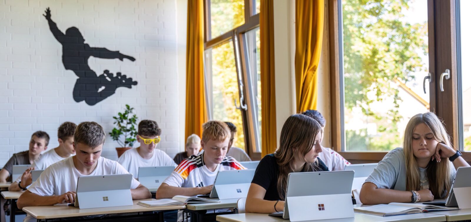 Schülerinnen und Schüler sitzen an Tischen im Klassenzimmer und arbeiten an Tabletts