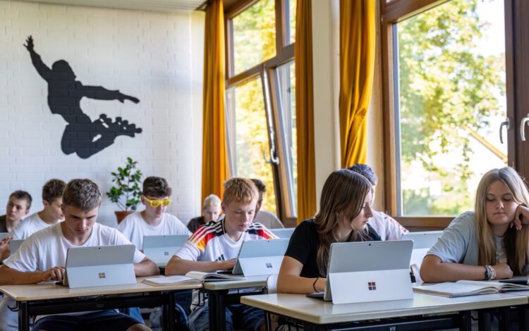 Schülerinnen und Schüler sitzen an Tischen im Klassenzimmer und arbeiten an Tabletts